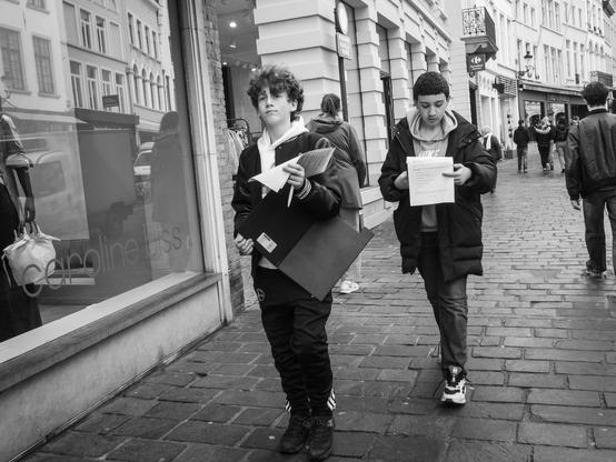 The image is a black and white photograph depicting two young individuals walking on a cobblestone street in Bruges, Belgium. The street is lined with buildings, and there are reflections visible in the window of a store named "Caroline Biss." The person on the left is holding a large black folder and a piece of paper, wearing a white hoodie, black pants, and dark shoes. The person on the right is reading a document, wearing a dark jacket with the word "NIKE" visible, jeans, and sneakers