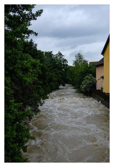 Foto im Hochformat. Blick auf einen reißenden Fluss, mehrere Meter breit. Das Wasser ist braun mit weißer Gischt. Links sind Bäume, rechts sind angeschnitten gelb verputzte Häuser. Davor sind Zäune und eine Mauer zum Wasser hin. Das obere ende der Mauer und das Wasser sind ca. einen Meter voneinander entfernt.