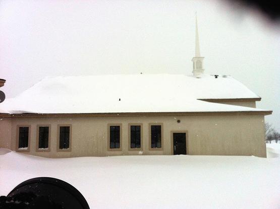 A dreary image taken on a very cloudy day in snowfall.  We are looking at a church, lengthwise, with its steeple to the right.  The roof is covered with snow, the church is a concrete colour, with dark, vertical windows.  Atop the roof peak we can see a grey blip. It is a snowy owl.