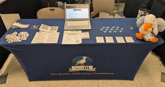 table with a blue cloth that contains SeaGL logo. on top of the table there is a seagull plushie bitting a French fry. there are stickers and posters with information on the conference.