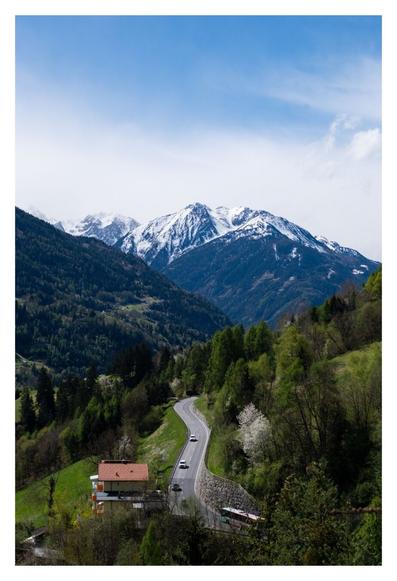 Foto im Hochformat. Blick über mehrere Berge. Im Vordergrund ein Berg mit Steigung links nach rechts, darauf wiesen, Bäume und eine Straße. Dahinter versetzt ein Berg mit Steigung rechts nach links, darauf ist ein Wald. Wiederum dahinter erhebt sich ein graues Gebirge, Richtung der Gipfel liegt Schnee. Die Straße vorne schlängelt sich Richtung Gebirge und wird befahren, drei Autos fahren bergab, ein Bus fährt bergauf. Der Himmel ist blau, rechts oben sind ein paar Wolken.