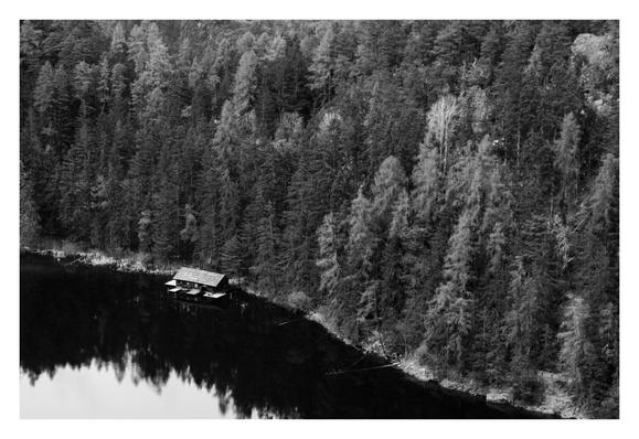 Schwarz-weiß Foto im Querformat. Teleaufnahme, ein Seeufer an einem Nadelwald von schräg oben fotografiert. Zwei Drittel des Bildes sind Nadelwald, die untere linke Ecke des Bildes ist der See. Am Ufer ist eine Hütte auf Stelzen im See. Diese liegt im schaffen der Bäume. Nur ganz links unten im Bild spiegelt sich der klar Himmel im Bild.