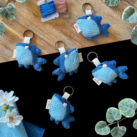 Five whale keychains photographed on a black and wooden background, surrounded with decorative plants and two shades of blue felt. 
The whales have a diamond-shaped body with bright blue bellies and dark blue backs and fins. Small black eye, pink blushes, and three clear blue lines are embroidered on their faces and bellies. 
On their heads are attached an ivory ribbon, linking them to small bronze-coloured key rings.
