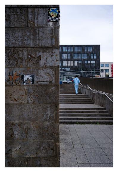 Foto im Hochformat. Die linkes Hälfte ist eine Wand in einer Großstadt. Sie ist gräulich braun, die einzelnen rechteckigen Steine sind zu erkennen. Verschiedene Aufkleber sind auf der Wand. Die rechte Hälfte des Bildes ist der Bereich neben der Wand. Vier Treppen mit flachen Bereichen dazwischen, rechts das Geländer. Eine Person in einem türkisfarbenen Trainingsanzug läuft die Treppe hoch. Weiter hinten sind verschieden hohe Gebäude, die dem bedeckten Himmel eine Treppenform geben.