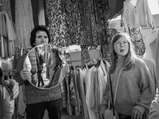 The image is a black and white photograph taken at a market in Bruges, Belgium. In the foreground, a man is holding a round mirror, reflecting the image of a woman with long hair, who is wearing glasses and a sweater. The man is wearing a textured sweater and dark pants. Behind them, various garments are hanging on racks, including dresses with floral and animal print patterns, as well as sweaters and jackets.