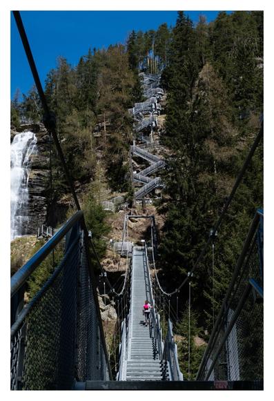 Foto im Hochformat. Blick auf eine riesige Treppe  aus Metall umgeben von Nadelbäumen. Im Vordergrund geht die Treppe geradeaus, auf beiden Seiten wird sie von dicken Kabeln gehalten. Eine einzelne Person mit rotem Rucksack läuft gerade empor. Weiter hinten geht die Treppe im zickzack nach oben. Links ist angeschnitten ein großer Wasserfall. Es ist sonnig.