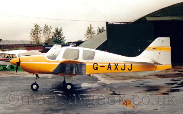 Side view of a white and yellow, single-engined light aircraft parked facing to the left.
The plane has white upper surfaces and a yellow lower half, with the registration "G-AXJJ" in black on the lower rear fuselage.
Black tarmac apron fills the foreground, with a black hangar with open doors in the background on the right, with another light aircraft on the left.
Trees and some other buildings are visible further back on the left, under grey skies.