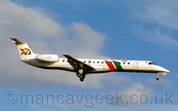 Side view of a white, twin engined jet airliner with the engine mounted on the sides of the rear fuselage, flying from left to right at a very low altitude with the undercarriage extended and the flaps deployed from the rear of the wing, suggesting it is about to land.
The plane is mostly white, with red and green bands running diagonally from the mid-cabin windows and under the body.
There are black "PGA" titles on the upper forward fuselage, with smaller "Portugalia Airlines" just below.
The white engine pods have small black "ERJ145" titles.
The plane has black "PGA" titles under half of a yellow sunburst in front of a diagonal red and green stripe.
Pale blue skies with fluffy white clouds fill the background.