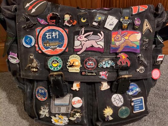 my bag at home, showing the complete collection (left to right, top to bottom):

three-arrows antifa
witch logo (out of shot)
anarchy heart
basic witch
jimquisition
antifa cogwheel
nezuko
moon tarot card
star tarot card
ace of cake
quark's bar
purple scryball
starfleet delta
federation
usg ishimura, glued over to hide a trans sylveon
ace espeon
gay eevee
witch logo
scp
cats and coffee
owl with cauldron
central perk
arrakis wormspotters
cult of the lamb
moon moth
d and d 20
raven with potion
doom floppy
umbrella corp
welsh dragon
"never again" abortion coathanger
usg ishimura
floppy disk
moomin
another moomin
i need space
AC/RC
men of letters
ff7 new game/continue
fox hound
zack
cloud
ATREIDES! ATREIDES! ATREIDES!
rpg action menu