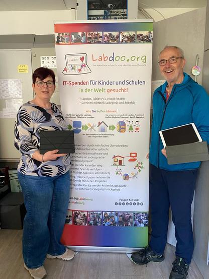 In einem Raum stehen die Leiterin des Kindergarten und ein Helfer von Labdoo vor einem Rollup von Labdoo. Beide halten jeweils ein Tablet in den Händen und schauen in die Kamera.