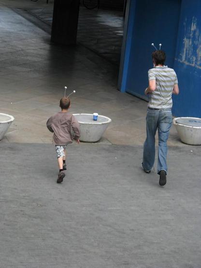 A photo of two people, young and adult, seen from the back running, wearing head boppers, outdoors on a concrete open space.