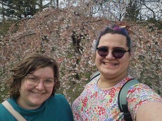 my partner and i taking a selfie in front of a tree blooming in pink, her with short slightly-curly hair and a turquoise overshirt, i with my coloured hair tied in a bun and a brightly-coloured floral dress.
