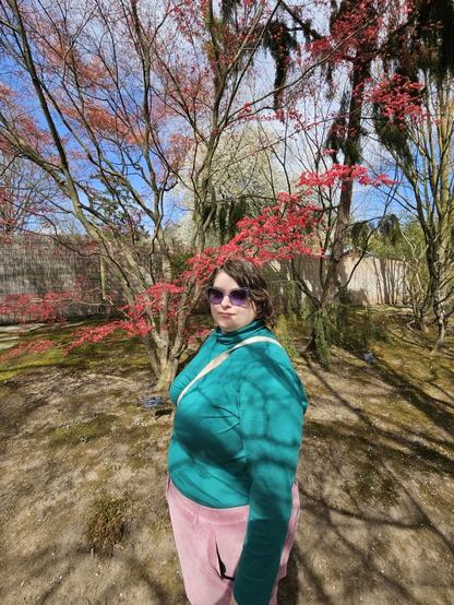 my partner with short slightly-curly hair and a turquoise-purple clothing combo that goes surprisingly well together, this time with sunglasses, standing in front of a tree blooming in strong red.