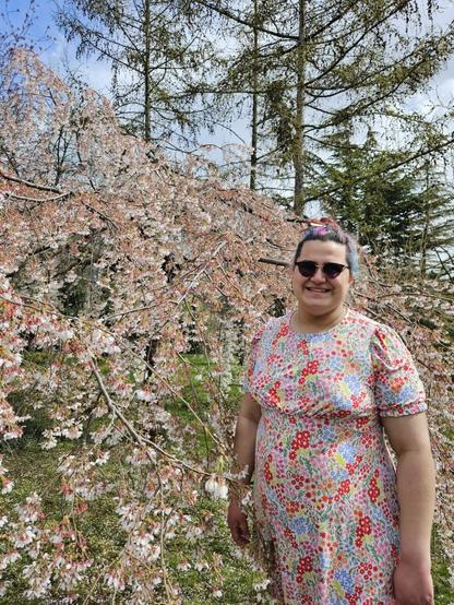 me standing next to a tree standing in pink, matching to my brightly-coloured floral dress. coloured hair tied in a bun, and i'm smiling.