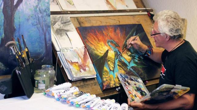 An artist with gray hair paints a colorful fantasy scene of a dragon at a cluttered workspace with brushes, paint tubes, and sketchbooks.