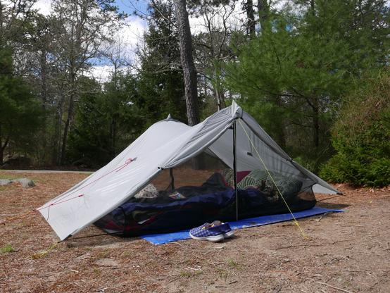 A two-person grey ultralight tent, of a tensioned-gable style, jam-packed with blankets and pillows, with trees in the background