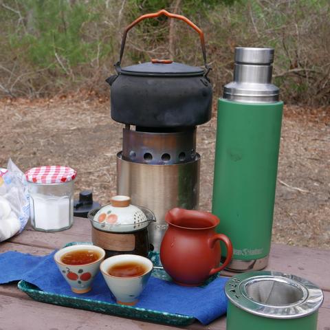 A tea set-up for out door hangs, consisting of a twig stove, a small kettle, a large thermos, a large glass gaiwan, a red clay fairness pitcher, two tea cups on a tea tray.