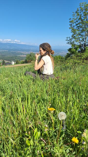 Woman in a green field drinking tea from a small brown tea cup, enjoying the view on a sunny day