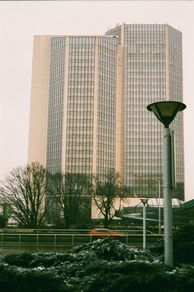 A photo of a modern glass and beige building with a motion blurred red car passing in front of it. The weather looks unpleasant