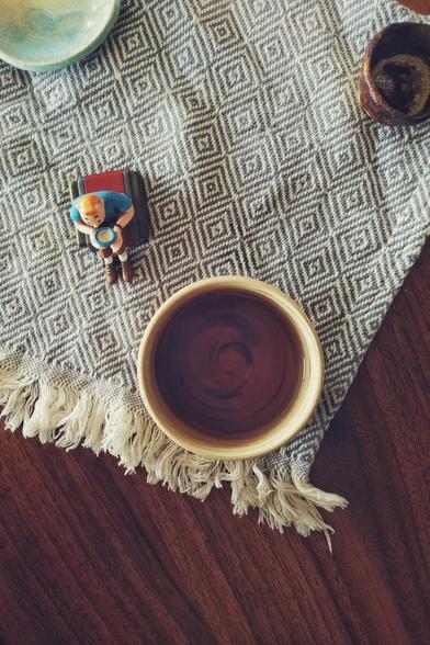 A vertical colour photograph of a small cup of tea full of dark tea. The cup is on a white and blue patterned cloth. Next to the cloth is a small Tintin figurine sitting and drinking tea. The table below the cloth is a dark brown. At the edges of the image there is a small decorative ceramic cup and a small decorative ceramic plate. both are cropped by the composition.