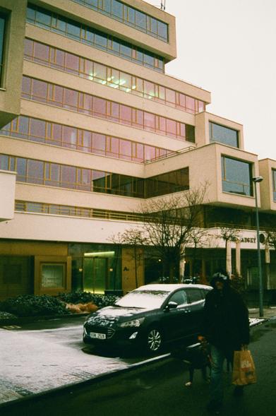A photo of a modern rectangular building with ochre walls and huge glass windows. The building is multi stored with each store being a different sized rectangle. There is also a dude and a dog in front of it