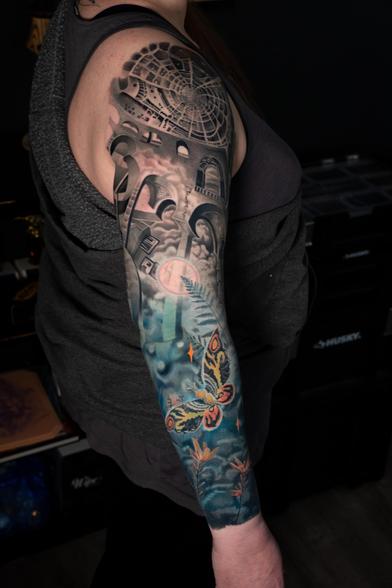 Photograph of a large tattoo sleeve on a woman’s right arm. The tattoo is in color realism style and features a moth, flowers and surrealism-style arches and staircases.