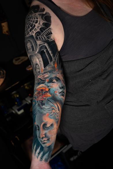 Photograph of a large tattoo sleeve on a woman’s right arm. The tattoo is in color realism style and features two statue-like women’s faces and surrealism-style arches and staircases.