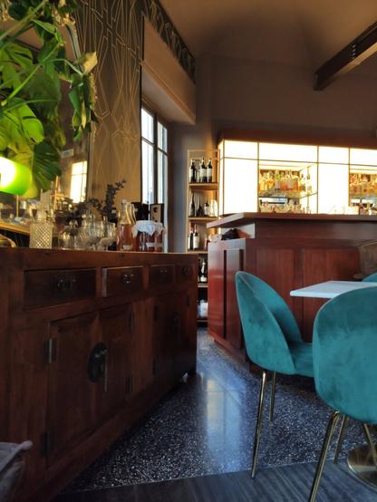 L'immagine ritrae l'interno di un cocktail bar. In primo piano sulla sinistra si nota un mobile in legno scuro in stile vintage, usato come credenza e postazione di servizio, con sopra caraffe, bottiglie di vetro e bicchieri, ben ordinati e valorizzati da un'illuminazione calda e accogliente.

A destra si intravedono due sedie in velluto verde petrolio con gambe dorate, abbinate a un tavolo bianco: un contrasto cromatico raffinato che suggerisce attenzione al design. Sullo sfondo, domina la scena un bancone bar in legno con retroilluminazione soffusa, che mette in risalto bottiglie e bicchieri disposti con cura su mensole di vetro. Accanto, una libreria a giorno con altre bottiglie completa l’allestimento.

Le superfici del pavimento, composte da materiali diversi (marmo e parquet), riflettono la luce e conferiscono ulteriore eleganza all’ambiente. Il decoro murale in stile Art Déco, con motivi geometrici dorati, contribuisce all’atmosfera sofisticata e curata del locale.
