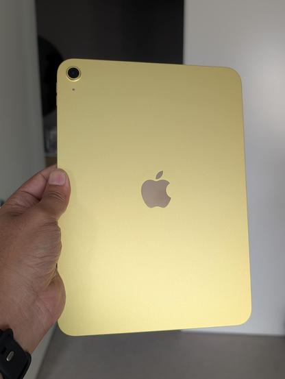 A person is holding a yellow Apple iPad with the Apple logo visible on the back. The iPad has a rear camera located at the top corner.