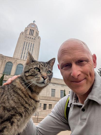 A bald white dude poses for a selfie with Cameron, the Capitol Cat, in front of the Nebraska State Capitol on a cool, cloudy, slightly wet morning.