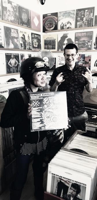 Monochrome medium shot in a cluttered record store. I'm in a black motorcycle helmet with goggles holding the Fanzui Xiangfa record, centered. Jason stands to the right, smiling and gesturing. Background is filled floor-to-ceiling with lined shelving covered in vinyl album covers. Light source visible above. Record bins visible on the bottom. Image uses a shallow depth of field with a slightly desaturated color palette.