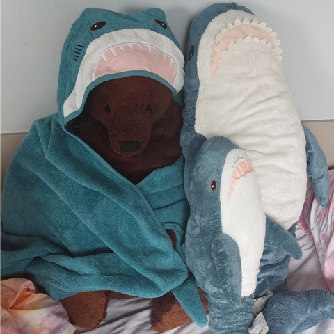 A cozy bedtime scene featuring two IKEA BLÅHAJ shark plushies taking care of a brown teddy bear. The teddy bear is snugly wrapped in what appears to be a BLÅHAJ-themed blanket or costume with the shark's face forming a hood over the bear's head. Beside them lies another blue BLÅHAJ shark on its side, creating a heartwarming cuddle puddle on pastel bedding. The sharks' signature details are visible - blue bodies, white bellies, and friendly open mouths with felt teeth. This sweet arrangement shows the BLÅHAJ sharks in their natural habitat - bringing comfort and companionship at bedtime, even to other stuffed animals.