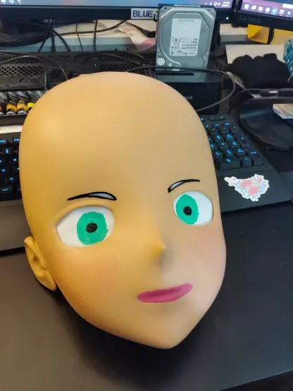 Photo of an in-progress Animegao Kigurimi mask without hair yet. It's painted a light fleshy color with bright green eyes, black eyebrows, and pink lips. A little bit of blush. It looks like an anime character in proportions but the paint job is whack
