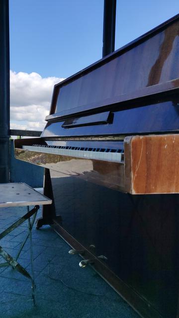 Ein Klavier und ein alter, feingliedriger Klapphocker stehen auf einer Steinterrasse. Am blauen Himmel direkt hinter ihnen ziehen Wolken vorbei und spiegeln sich im schimmernden Klavierlack.