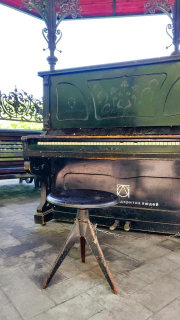 Unter einem Pavillon mit tiefrotem Dach und verschnörkelten gusseisernen Streben steht ein altes mattschwarzes Piano. Ein runder, drehbarer Klavierhocker ist daran angekettet. Oberhalb der Klavier-Pedale sind ein Logo aus geometrisch ineinander verschachtelten Formen und der teilweise vom Hocker verdeckte Schriftzug покоління відкритих людей erkennbar.