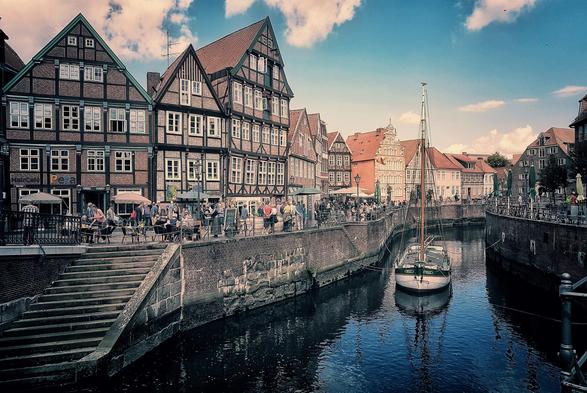 A view of the German town of Stade, showing a canal lined with historic brick buildings and a small boat docked in the water under a partly cloudy sky.