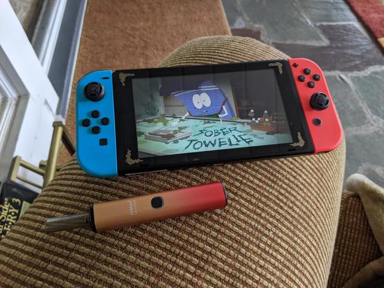 Photo of my Nintendo Switch with South Park: The Fractured but Whole loaded. The Sober Towelie mission is currently onscreen. Next to the Switch is my IMax V3 Nano vaporizer with Ms Jackson cannabis flower loaded.