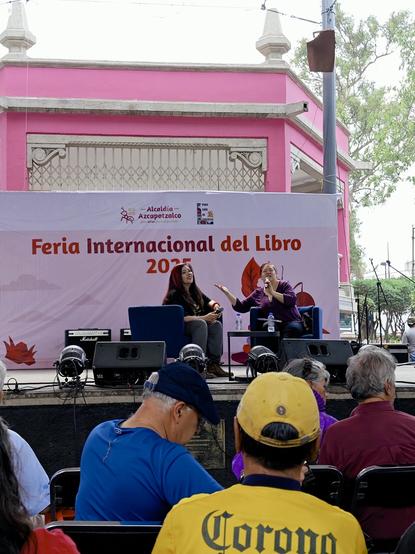 En un escenario levantado delante de un kiosco pintado de rosa, una mujer de cabello oscuro –la escritora Eve Gil– es presentada por otra de cabello rojo: la escritora Raquel Castro. Esta sostiene un micrófono y ambas están sentadas en sillones azules.