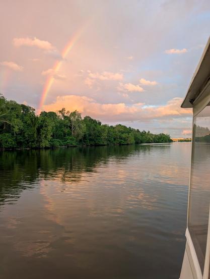 Rainbow over green tree lined bank and stern of boat in river reflecting in water