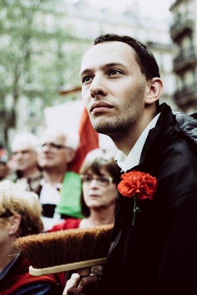 Un jeune homme écoute les discours avec attention, il a un balais à la main et un oeillet rouge pimpant à la boutonnière de sa veste noire.