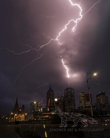 A dramatic lightning storm crackles over Melbourne’s city skyline at night. Brilliant white bolts stretch across the purple-hued sky in jagged, electrifying lines. Below, the city buildings glow with interior lights, including the illuminated spire of St. Paul’s Cathedral and the towers of Federation Square and the CBD. The contrast between natural fury and urban stillness creates a cinematic, high-energy moment.