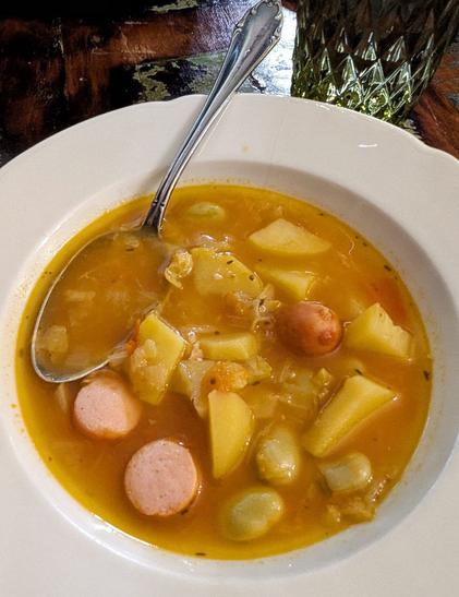 Dunkler Tisch, weißer Suppenteller mit rotgelber Gemüsesuppe darin. Es sind Kartoffelstücke, Kohl, dicke Bohnen und Würstchenscheiben erkennbar.  Ein silberner Löffel liegt im Teller.