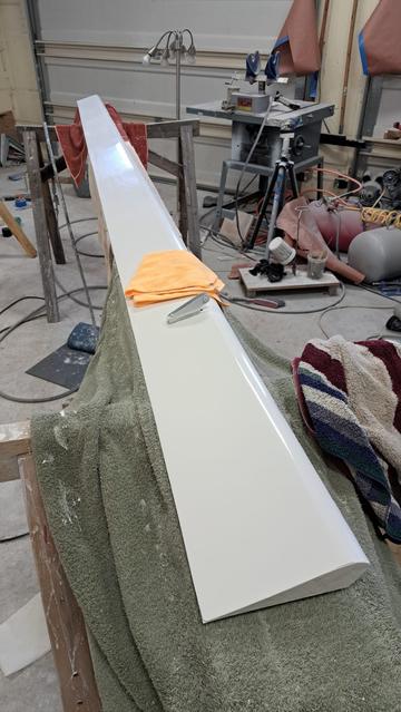 The white flaperon, 12 feet long, sits on a padded work surface inside a garage workshop. Its white paint has been buffed and polished. An orange/yellow polishing rag sits on the flaperon. In front of the polishing rag is the drive horn that will be connected to a push-pull rod that is part of the flight control mechanical linkage.