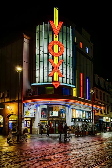 Facade of splendid Art Deco style Cine vox cinema in Strasbourg at night, illumitated by its colourful red, blue and yellow neons. Tiny silhouettes of 4 people, gathering before the building for an evening screening, are visible.
