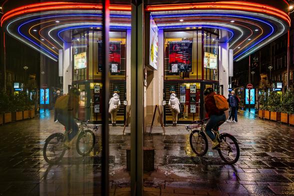 Symmetrical reflection shot in a shop window of the facade of splendid Art Deco style Cine vox cinema in Strasbourg at night, illumitated by its colourful red, blue and yellow neons. A cyclist is visible in the foreground.
