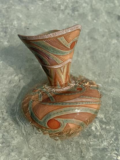 A large ceramic vase (the same one as in the original post) with a voluminous, bottom portion, narrow waist and flared neck, at the beach washed over by clear blue ocean water. The vase is intricately decorated with earth tones, and a light aqua glaze. Patterns swirl around the vase, just as the waves and water do. 

This photo was taken on the same day as the photo in the original post. The angle of the vase in the water is different and the lighting is slightly different, as well. The vase stands more upright and sunlight dances in the water around the vessel.