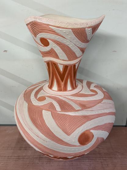 The same vase in the phase after bisque firing and glazing but before the final glaze firing. Instead of rich, deep earth tones and aqua, we see pink and white tones that will transform in the kiln. The piece sits on a wood shelf with a pale blue-green wall in the background.