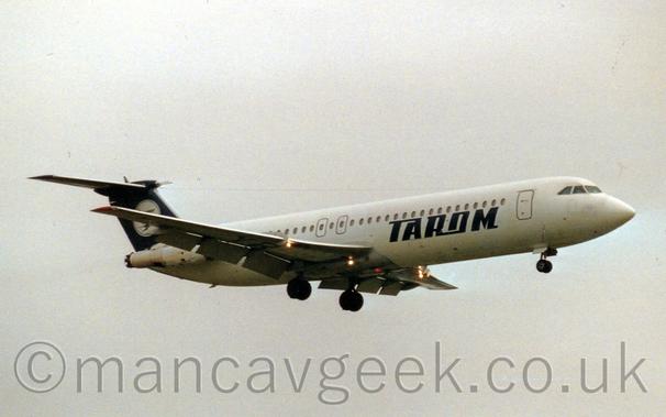 Side view of a white, twin engined jet airliner with the engines mounted on the sides of the rear fuselage, flying from left to right at a very low altitude, with undercarriage lowered and flaps extended from behind the wings, suggesting it is about to land.
There are dark blue "Tarom" on the lower forward fuselage.
The tail is dark blue, with a white circle containing a dark blue flying bird.
Several bright lights are shining forward from under the wings.
Flat grey sky fills the rest of the frame.