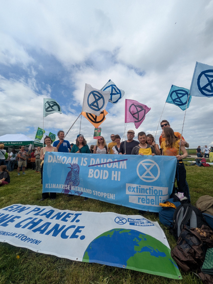 Aktivistengruppe mit bunten XR-Fahnen und hellblauem XR-Banner
"DAHOAM IS BOID HI"
Davor am Boden liegt ein Banner
"EIN PLANET EINE CHANCE"
