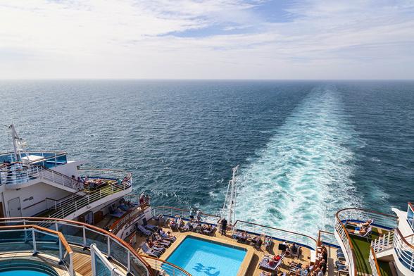 Taken from a high deck at the aft of a cruise ship we're looking down diagonally across the stepped decks below us that border the aft pool of the ship, a square pool with space around it for a couple of dozen sun loungers, most occupied. A handful of loungers can also be seen on some of the terraced deck space around and above it and just in shot to our left is the edge of another small, circular pool or hot tub here in this location. The sky is bright but with a fair amount of thin sheets of hazy cloud that's diffusing the sunshine, but there's still plenty of that to warm this spot on the vessel. The sea is calm, a deep turquoise colour, and with a long wake from the ship tapering off towards the horizon as a churned, foamy trail on the sea's surface.